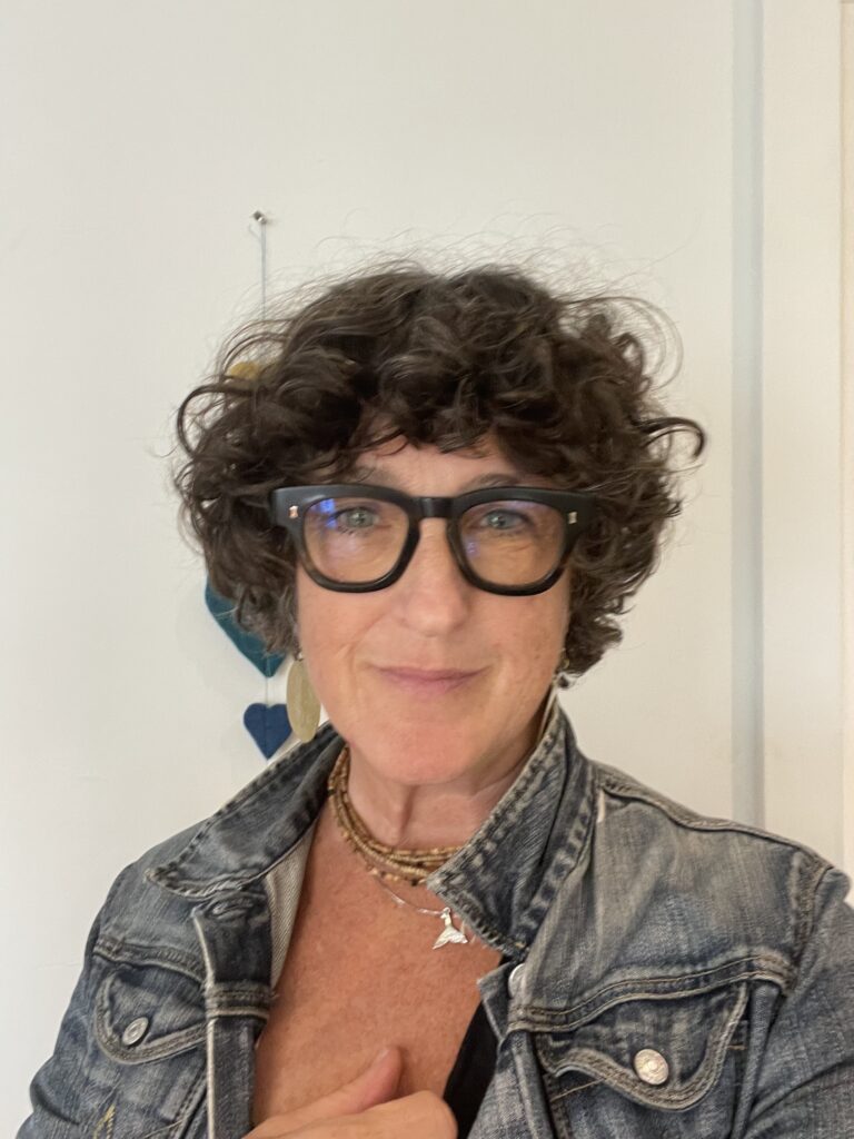 Jo Tooth - short dark hair with glasses, denim jacket with hand on hear -  founder of Dhama Sun, smiling in Cornwall — conscious living coach