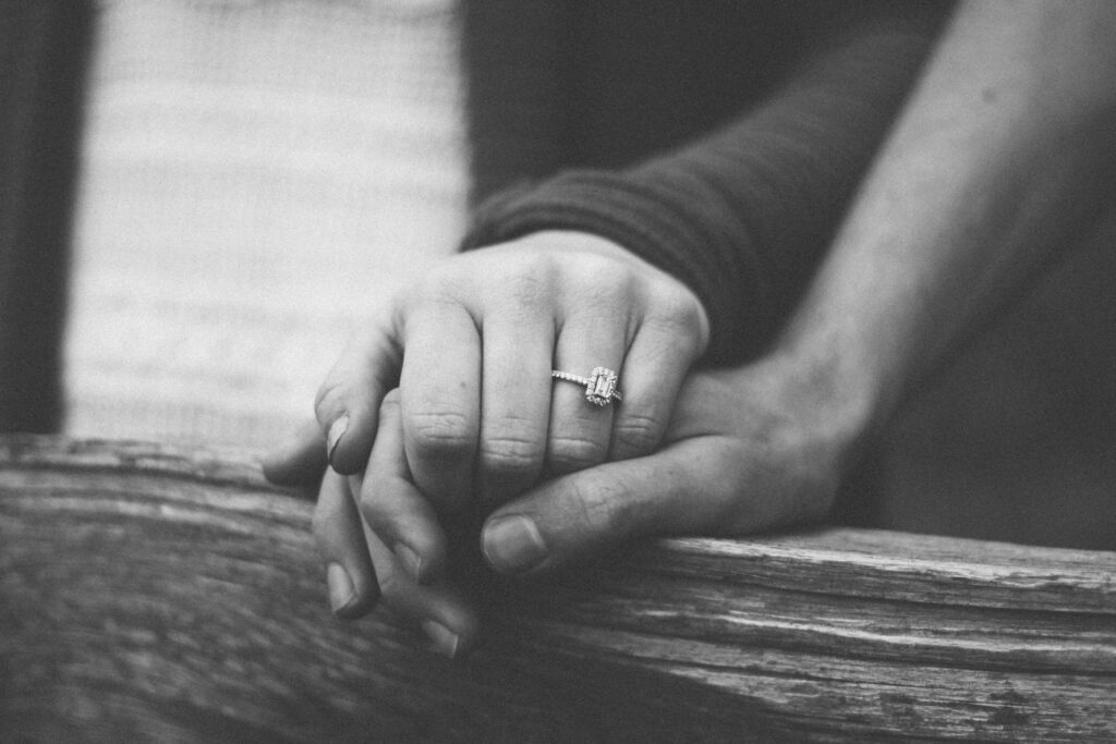 Black and white picture showing two hands, one has a diamond engagement ring and the hands are resting on a wooden ledge - depicts togetherness, compassion, love